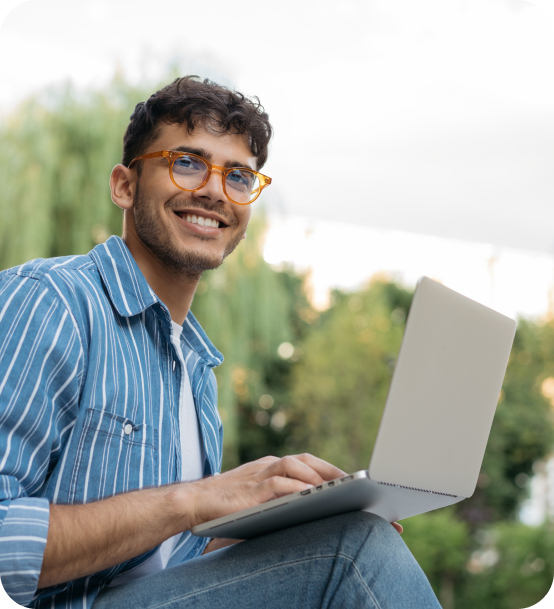 Image of young man with glasses smiling while sitting outside holding a laptop.
