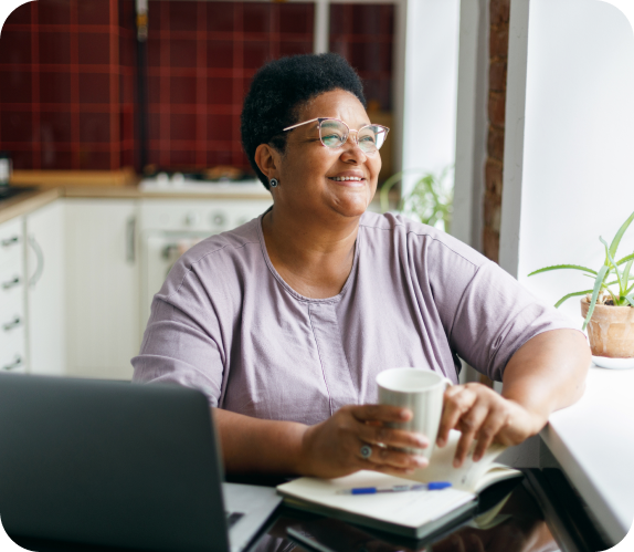 Image of older black woman smiling and looking out the window while sitting at her desk at home in front of a laptop and drinking coffee or tea.